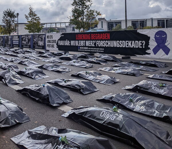 Demonstration mit Banner 'Lebendig begraben', davor mehrere Reihen von Leichensäcken mit blauen Rosen darauf