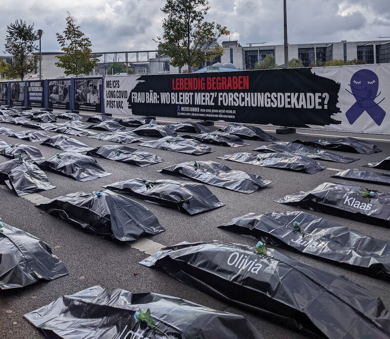 Demonstration mit Banner 'Lebendig begraben', davor mehrere Reihen von Leichensäcken mit blauen Rosen darauf