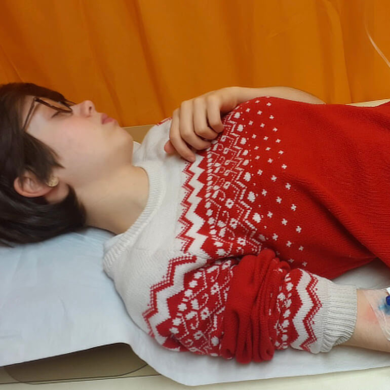 Juliane liegt mit geschlossenen Augen auf dem Rücken, im Arm eine Infusion