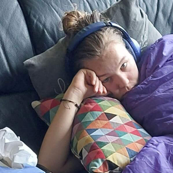 Leonie liegt mit Kissen, Decke und Kopfhörern auf dem Sofa und schaut traurig in die Kamera