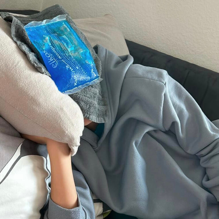 Levi mit ME/CFS und Handtuch und Kühlpack auf dem Kopf auf dem Sofa liegend