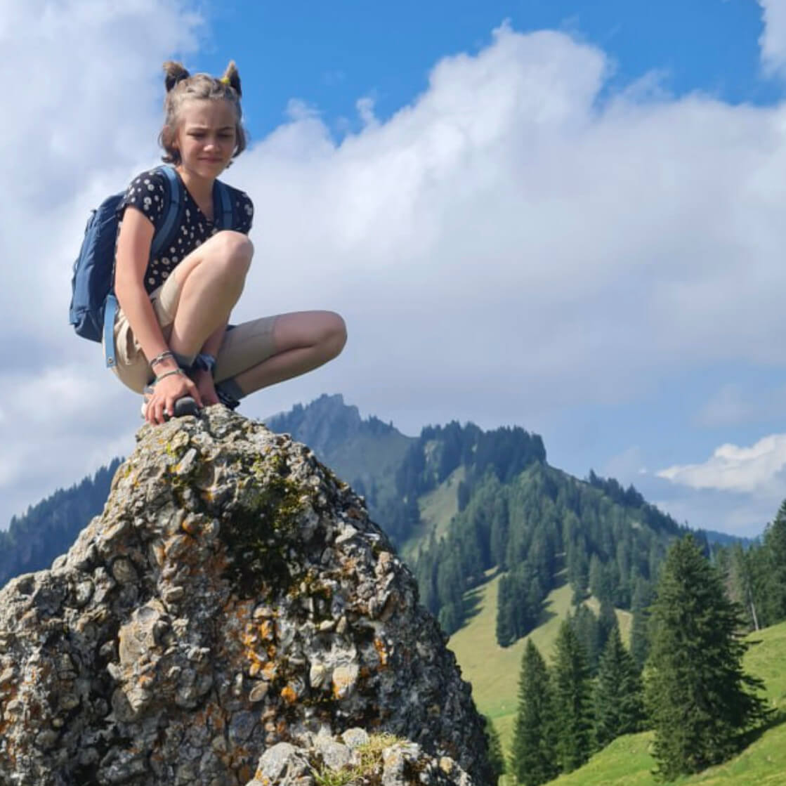 Lina vor ME/CFS. Sie hockt auf einem erhöhten Stein, im Hintergrund eine Berglandschaft
