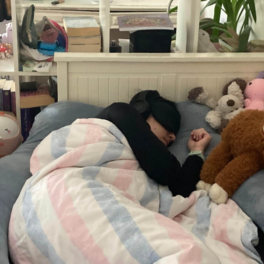 Ronja liegt mit Augenmaske und Gehörschutz im Bett, um sie herum Bücher und Kuscheltiere